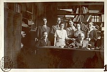 Photographie en noir et blanc d'un groupe de quatre femmes et quatre hommes derrière un bureau et devant une étagère avec des livres