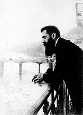 A long-bearded man in his early forties leaning over a railing with a bridge in the background. Dressed in a black overcoat, he gazes blankly into the distance with his hands clasped.