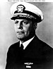 Head and shoulders of a middle aged white man wearing a white peaked cap with a black visor and a dark jacket over a white shirt and dark tie. On his left breast is a winged pin and a single ribbon bar.