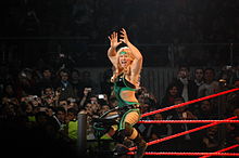 A blonde woman sits on the turnbuckle of a wrestling ring, stradding the ring post, and facing the crowd while posing with her hands in the air. She is wearing a green and black catsuit with the sides of the torso removed to bare part of her midriff.