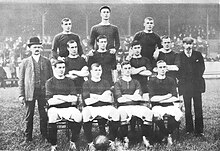 A black-and-white photograph of a football team lining up before a match. Four players, wearing dark shirts, light shorts and dark socks, are seated. Four more players are standing immediately behind them, and three more are standing on a higher level on the back row. Two men in suits are standing on either side of the players.