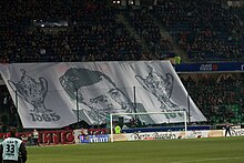 Photographie montrant un portrait peint sur un grand linge, porté par des personnes dans une tribune de stade