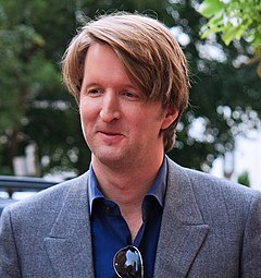 A man in a gray suit with a blue shirt looks down to his right while smiling.