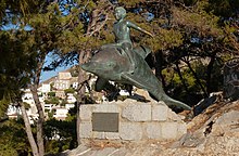 Bronzestatue, die einen auf einem Delphin reitenden Jungen zeigt. Im Hintergrund Kiefern und der Hafenort Hydra.