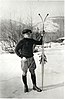 A young man standing beside skis in front of a hilly, snowy background