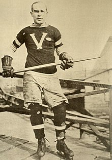 A man poses for a photo wearing skates and an ice hockey sweater, and holding a stick in his hands.