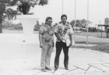 Deux hommes en chemise debout en extérieur, avec un grand collier de fleurs autour du cou