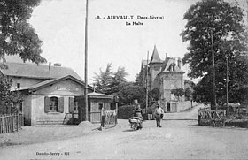 Carte postale noir et blanc, à gauche le bâtiment de la halte, au centre le passage à niveau traversé par une femme ayant une brouette et un homme qui l'accompagne, en arrière plan une qui se sépare en deux avec au centre une grande villa, à droite une barrière et un grand arbre.