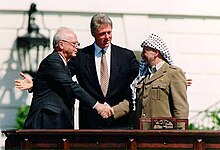 A smiling Arafat, donning traditional Arab headdress, shakes the hand of a stolid Rabin as Clinton stands tall behind and center with open arms toward the two leaders.