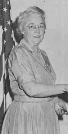 An older white woman with white hair, wearing glasses and a light-colored dress in front of an American flag