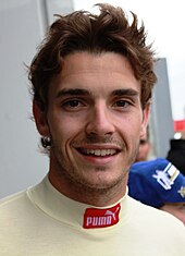 Man in his early twenties with a head full of hair and smiling. He is wearing white racing overalls.