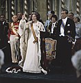 Une femme en robe blanche et portant un manteau d'hermine et un cordon orange, lève la main droite en étant debout devant un trône, alors qu'elle est entourée de personnes en uniformes et costumes d'apparat.