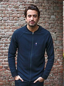 Dotan wearing a dark blue zip-up jacket and dark pants, hands partly inserted in pant pockets, standing in front of a brick wall, grinning slightly at camera