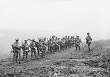 A line of soldiers in battle equipment face another soldier who is addressing them on a gentle slope. Behind them, smoke or fog obscures the rest of the terrain.