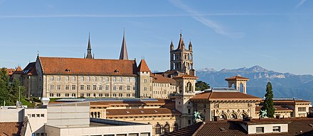 Paysage urbain vu des toits ; un bâtiment gothique imposant se distingue en arrière-plan sur fond de montagnes.