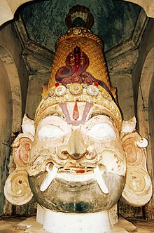 A big wooden male head with a moustache, bushy brows, Vaishnava tilak, fangs protruding from the end of the mouth, big eyes and ears and wearing a conical crown with a cobra on its front and another five-headed cobra is emerging from behind