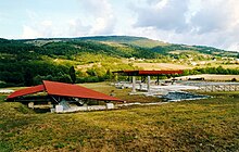 Vestiges avec colonnes et murs, partiellement recouvert par une toiture moderne de protection avec une colline, des champs et des bois en arrière-plan.