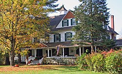 A large white house with green trim mostly concealed by trees in front, one of which is beginning to change to autumn color