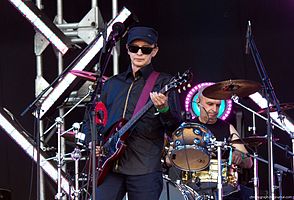 Edmund Shklyarskiy of Picnic wearing a dark shirt, pants, and cap, with black sunglasses, playing electric guitar on an outdoor stage, with drummer behind him