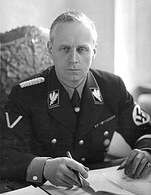 Portrait of a middle-aged man with short grey hair and a stern expression. He wears a dark military uniform, with a swastika on one arm. He is seated with his hands on a table with several papers on it, holding a pen.