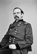 Black and white photo shows a seated man with a moustache and goatee. He wears a dark military uniform with the two stars of a major general on the shoulder tabs.