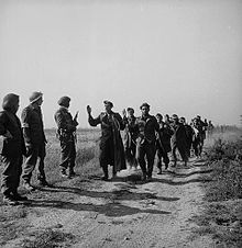 Photo en noir et blanc de militaires marchant sur un sentier avec les mains en l'air et de d'autres militaires se tenant debout à côté du sentier les regardant