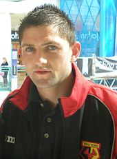 A young man wearing a black shirt and shorts, standing on a grass field.