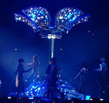 A bluish image of a pillar on a stage. It is flanked by women in horse-riding gear. From both the ends of the pillar hang a number of plates, the inside of which is lit by blue lights. One of the bottom plates has a number of stairs on which a similar horse-riding gear dressed lady, with her back to the image, is coming down.