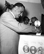 A man wearing a suit inserting a piece of paper into a box. He is being photographed by cameramen