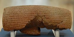 Front view of a barrel-shaped clay cylinder resting on a stand. The cylinder is covered with lines of cuneiform text