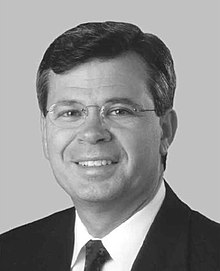 A dark-haired man in his forties wearing glasses and a suit. He is smiling and facing left.