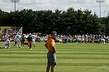 Rear 3/4 photograph of Angelo wearing an orange t-shirt and grey shorts standing on the practice field at Chicago Bears training camp