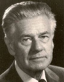 Formal sepia portrait of man in suit and tie, with strong features and greying hair brushed back.