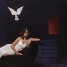 PinkPantheress in a white dress reclining on a dark platform as a white dove flies over her