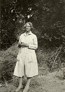 Woman standing in front of plants, wearing a long coat and glasses