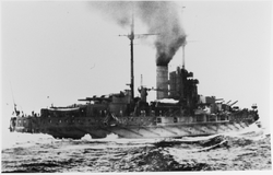 A large battleship steaming away through the water at high speed. Its stern can be seen in the foreground with water hitting the sides of the ship. Smoke can be seen billowing out of the funnels of the ship.