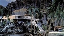 A luxury villa on top of a volcanic rock formation, with palm trees in front of it, a rock face behind, flights of steps leading down from it and a swimming pool to one side.