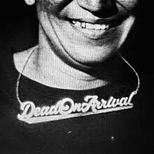 A monochrome image of a person smiling with a shirt and a necklace reading "Dead On Arrival" with the words underlined.