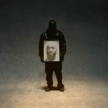 Ty Dolla Sign completely covered in a black leather jacket and mask. He is holding a picture of his brother "Big TC" and standing in the center of a graduated color background going from tan, to cerulean, to black at the top.