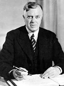 A middle-aged man in a dark suit and stripped tie sits behind a desk. A book is open on the desk and he holds a pen in his right hand.