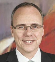 Colour publicity shot of a clean-shaven, smiling, bespectacled man with receding, greying hair.