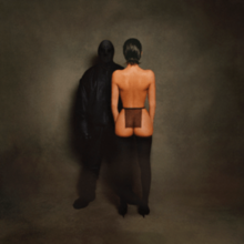 A photograph of Ye completely obscured by all-black clothing (including an opaque head covering) standing next to his wife, Bianca Censori, with her back turned, wearing stockings and a sheer black cloth around her waist