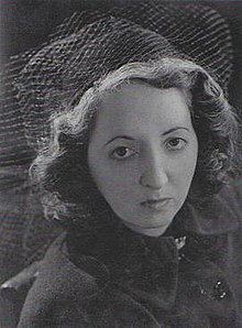 A young woman looks into the camera with a serious expression, her short, wavy hair topped by a hat with veiling