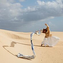 A red headed woman wearing a beige shirt and white skirt stands in the middle of a desert with the wind blowing her arms and a large drapery that flows from her hand.