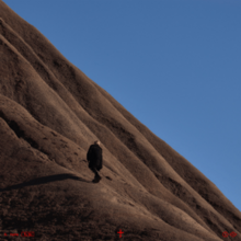 Irama with his back to the camera walking along a very steep hill with a blue sky in the background