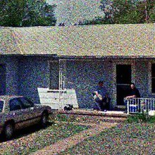 A grainy image of two individuals sitting on the front porch of their modest home; a car is parked in the driveway.